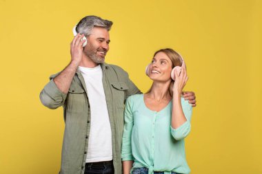 Kulaklıklı mutlu orta yaşlı çift müzik dinliyor, sarılıyor ve birbirlerine sarı arka planda bakıyorlar. Pozitif kadın ve erkek favori müzikleri seviyor
