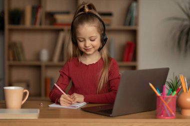 E-Learning. Kid Girl Learning Online Using Laptop Computer Doing Homework Wearing Headset Sitting At Desk At Home. Remote Education. Educational Internet Service Concept