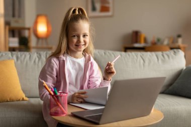 Dizüstü bilgisayarın başındaki kız, evde ödev yaparken kameraya gülümseyen notlar alıyor. Modern Eğitim ve Eğitimsel İnternet Hizmetleri Kavramı