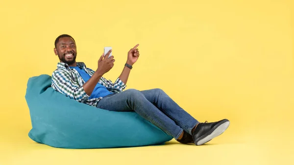 Happy Black Guy Using Phone Pointing Finger Aside Sitting Beanbag — Fotografia de Stock
