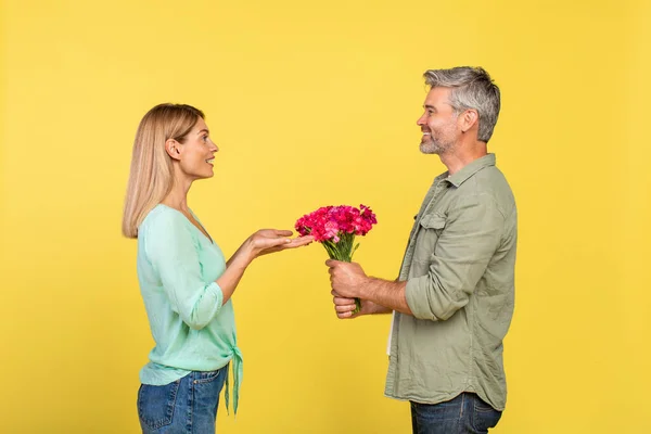 Happy Mature Man Presenting Flowers His Wife Yellow Studio Background — Stockfoto