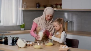 Little helper. Cute girl helping her muslim mother in hijab to cook breakfast, whisking eggs with milk in bowl and smiling, free space