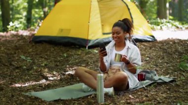 Internet connection anywhere. Young carefree african american woman tourist web surfing on smartphone, resting near tent at woodland, tracking shot, slow motion