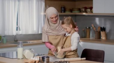 Domestic baking. Adorable little girl helping her mother to knead dough at kitchen, proud loving woman wearing hijab watching her kid, slow motion, free space