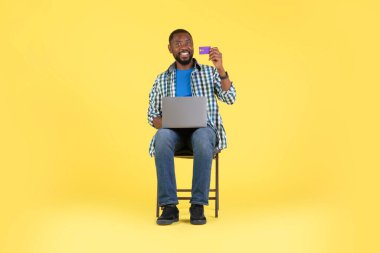 Smiling Black Man Shopping Online Using Laptop Holding Credit Card Sitting In Chair Over Yellow Studio Background. Internet Banking And Ecommerce Concept