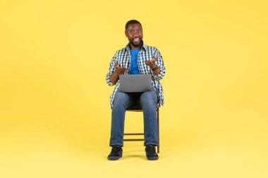 African American Man Video Calling Using Laptop Computer Talking Looking At Camera Sitting Over Yellow Studio Background. Modern Distance Communication Concept