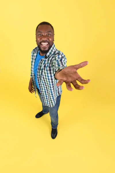 Happy African American Male Stretching Hand To Camera Holding And Showing Invisible Object Standing Over Yellow Background In Studio. Guy Advertising Product. Vertical Shot