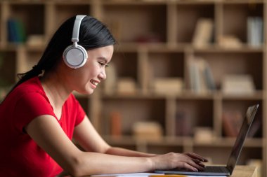 Cheerful young korean female with down syndrome in headphones watching online lesson in room interior, profile. Tutoring, homework, education at home, study and social distancing during covid-19