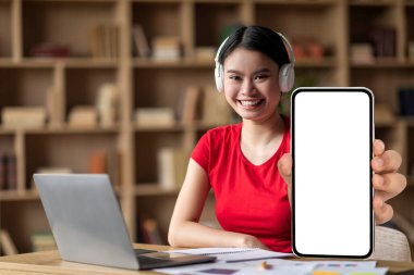 Smiling young asian woman with down syndrome in headphones with laptop shows phone with blank screen in room interior. Modern technology for education and knowledge at home, study remote, ad and app