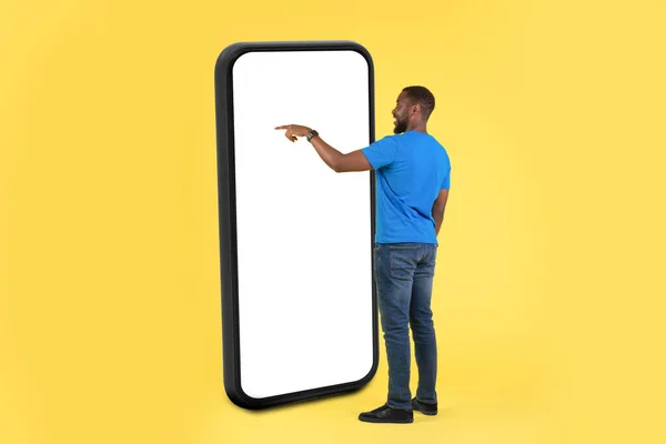 Side View Of African American Man Using Phone With Huge Blank Screen Standing Over Yellow Studio Background. Guy Using New Application. Gadgets And Technology. Mockup