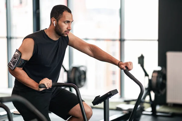 Sporty Young Black Man Using Elliptical Bike Machine While Training At Gym, African American Male Athlete Exercising With Modern Fitness Equipment In Sport Club, Enjoying Cardio Workout, Free Space