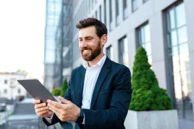Smiling millennial european man with beard in suit chatting on tablet on balcony of modern office building, outdoor. Ceo manager use gadget for develop marketing strategy, business remotely and app