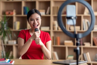 Cheerful pretty young japanese female superstar singing at microphone, shooting blog on smartphone in room interior. Hobbies and modern technology, karaoke and entertainment during covid-19 outbreak