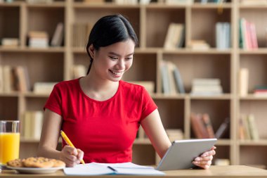 Cheerful pretty teenager japanese female student studying with tablet, enjoy home education remote in cabinet interior. Modern technology for study, online lesson and snacks during covid-19 outbreak