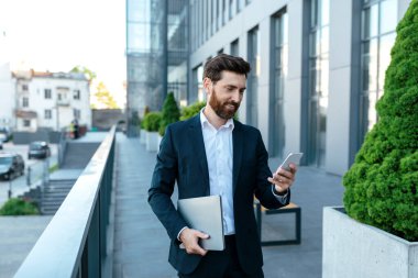 Smiling confident young businessman with beard in suit with laptop typing on smartphone near modern office building, outdoor. Manager and successful business, work with device remotely, chat in blog
