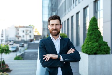 Successful business and work. Happy confident millennial handsome businessman with beard in suit with crossed arms on chest near modern office building, outdoor. Professional, ceo manager and boss