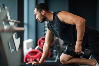 Young African American Sportsman Training With Dumbbell At Gym Interior, Side View Shot Of Black Male Athlete Lifting Heavy Barbell With One Hand, Working Out Arm Muscles, Free Space