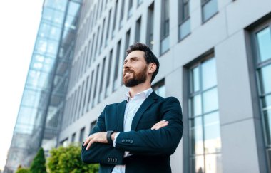 Smiling confident handsome young businessman with beard in suit with crossed arms on his chest looks at empty space near office building, outdoor. Leadership, ceo manager and boss lifestyle concept
