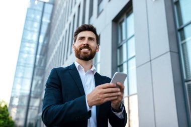 Gadget for business remotely. Cheerful confident millennial european man manager with beard in suit chatting on smartphone and looking at free space near modern office building, outdoor. Ad and offer