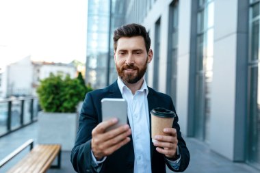 App for business and work. Confident millennial attractive european businessman with beard in suit with coffee takeaway, typing on smartphone near modern office building. Device for meeting remotely