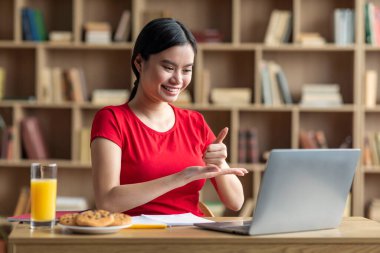 Glad teen korean female student studying, showing thumb up gesture with hands at laptop in room interior. Education at home, video call and meeting, snacks and modern technology due covid-19 outbreak