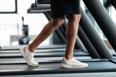 Jogging Workout. Cropped Shot Of Black Male Training On Treadmill At Gym, African American Man Legs In White Sneakers Shoes Running On Path In Fitness Club, Side View With Selective Focus
