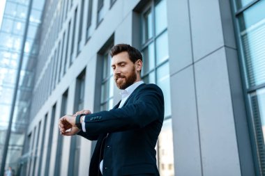 Smiling confident handsome young european male with beard in suit looks at watch, waits client near modern office building. Successful businessman, ceo manager and boss, business meeting and work