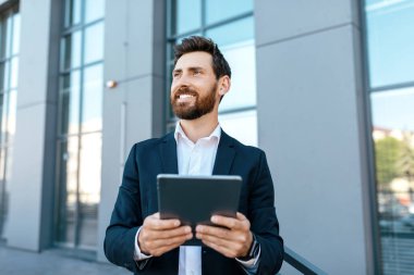 Happy confident millennial european man with beard in suit use tablet, look at empty space near modern office building, outdoor. Successful business, new app and video meeting with ceo manager, boss