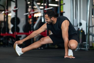 Sporty Young Black Man Stretching Leg Muscles While Training At Gym, Smiling African American Male Athlete Warming Up Before Workout In Modern Sport Club, Motivated Guy Exercising Indoors, Copy Space