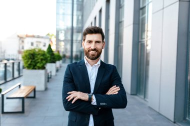 Leadership, ceo manager and boss. Smiling confident young attractive european man with beard in suit with crossed arms on chest near modern office building, outdoor. Work and business, ad and offer