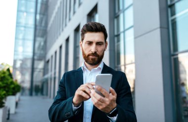 Serious confident millennial caucasian businessman with beard in suit typing on smartphone near office building, outdoor. Ceo manager and boss use gadget for meeting, business online, new app for work