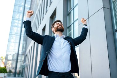 Cheerful attractive young caucasian businessman with beard in suit raises his hands up and rejoices to victory near modern office building, outdoor. Ceo manager, boss enjoy successful business and win