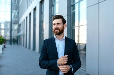 Smiling confident millennial attractive caucasian businessman with beard in suit looks at free space stands near modern office building, outdoor. Successful ceo manager and boss, business in city