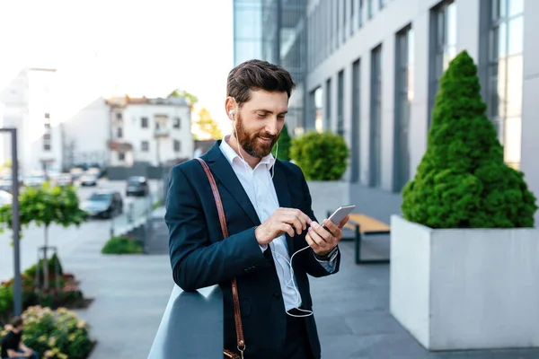 Smiling Millennial Caucasian Businessman Beard Suit Headphones Typing Phone Listen — Zdjęcie stockowe
