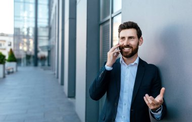 Cheerful confident millennial european businessman with beard in suit calling by smartphone near modern office building, outdoor. Ceo manager, boss has meeting remotely with client for business