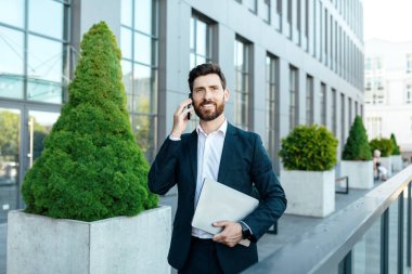 Busy smiling confident young caucasian businessman with beard in suit with laptop, call by smartphone near modern office building, outdoor. Ceo manager and successful business, work with technology