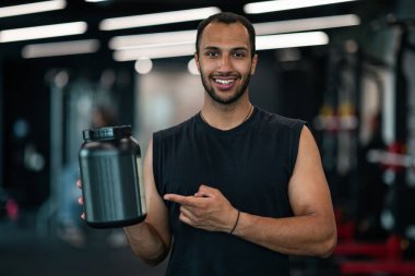 Handsome Black Male Athlete Advertising Fitness Supplements While Posing At Gym Interior, Smiling Young African American Sportsman Holding And Pointing At Dark Container With Whey Protein Powder
