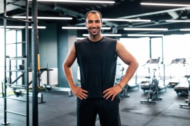 Portrait Of Handsome Black Personal Trainer Posing In Fitness Club Interior, Confident African American Sport Couch Standing With Hands On Waist And Smiling At Camera, Ready For Workout, Copy Space
