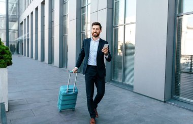 Smiling millennial european businessman with beard in suit and smartphone goes to airport with suitcase near modern office building, outdoor. Manager at business trip, work, journey and technology