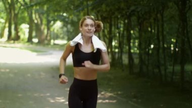 Active morning habits. Sporty middle aged woman athlete wearing earbuds running in public park with towel on shoulders, follow shot, slow motion