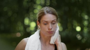 Exhausting sport training. Close up portrait of tired woman athlete wearing earbuds wiping her face with towel, feeling overworked after workout outdoor, slow motion