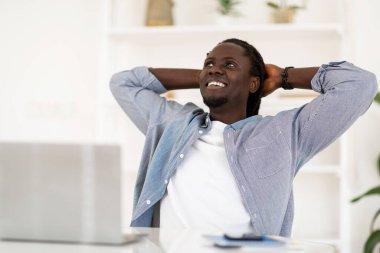 Relaxed Black Male Freelancer Leaning Back In Chair, Resting After Online Work With Laptop In Home Office, Dreamy Young African American Man Sitting At Desk With Hands Behind Head And Looking Aside