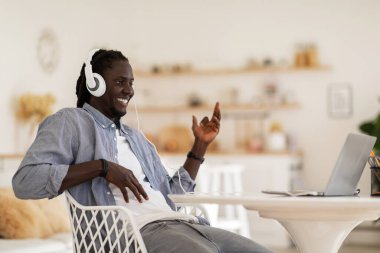 Cheerful Black Guy Wearing Headphones Playing Virtual Guitar While Listening Music On Laptop, Positive African American Man Having Fun While Working Remotely From Home Office, Copy Space
