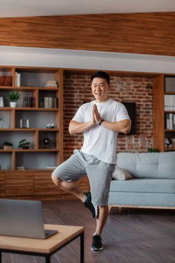 Excited asian mature man in sportswear attending online yoga class, standing in tree pose in front of laptop and smiling, home interior, copy space, full length shot