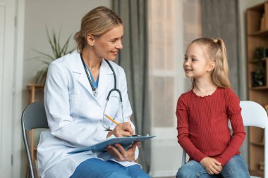 Pleasant female pediatrician talking to pretty schoolgirl patient and writing down notes in medical history paper during home calling visit. Healthcare concept