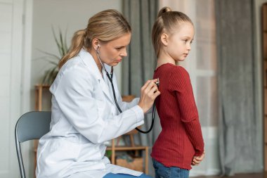 Female pediatrician listening to heartbeat and lungs by applying stethoscope to back of little girl. Doctor came to home to examine the child