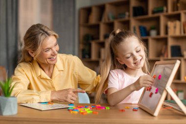 Happy female teacher and little girl studying, child making works on flip chart, sitting at desk in office. Positive private tutor having lesson with pupil. Education concept