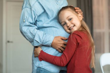 Pretty little girl hugging her pregnant mother big belly and smiling at camera, kid bonding with expecting mom, looking forward to become older sister