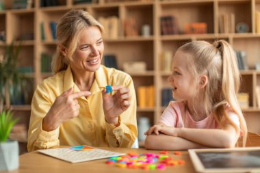 Happy woman speech therapist teaching little girl with pronounciation deffects to say sound R during personal training at classroom