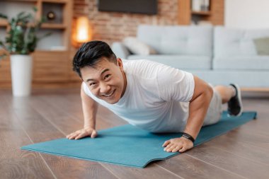 Strong asian mature man making strength workout, standing in plank pose or doing push ups at home on yoga mat. Happy male training his body, leading active lifestyle